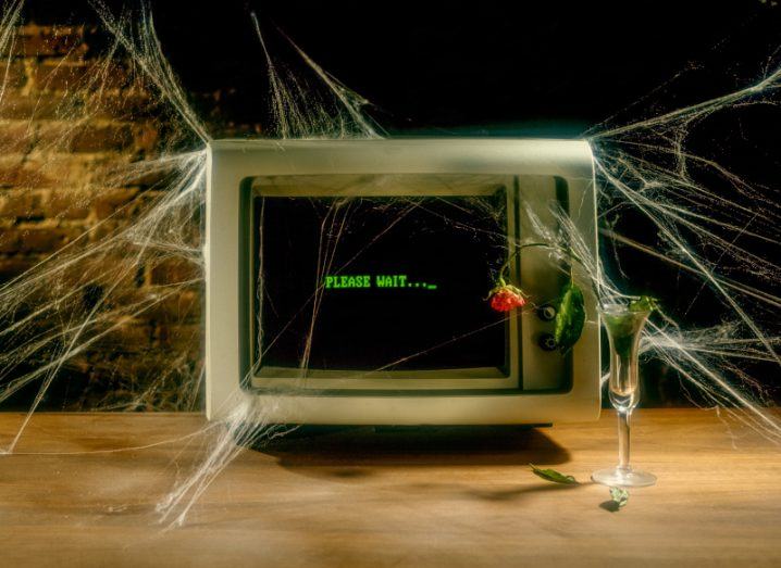 An old CRT computer sits on a table covered in cobwebs with a wilting flower sitting next to it. There is green text on the screen saying "please wait".