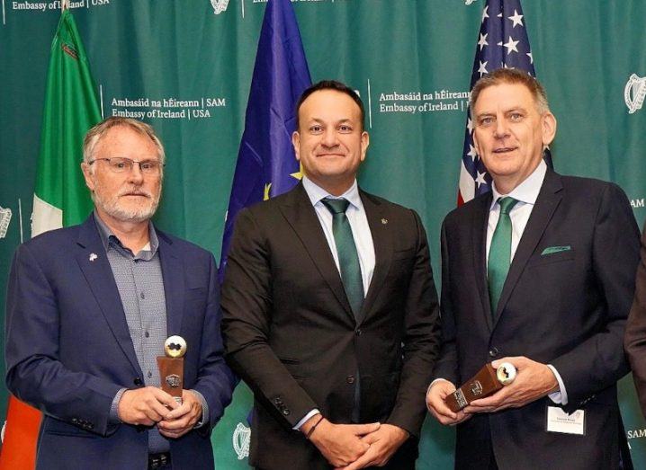 John Hartentt and Eamonn Keogh stand on either side of Taoiseach Leo Varadkar holding their St Patrick's Day medal in front of a green backdrop with Irish and US flags.