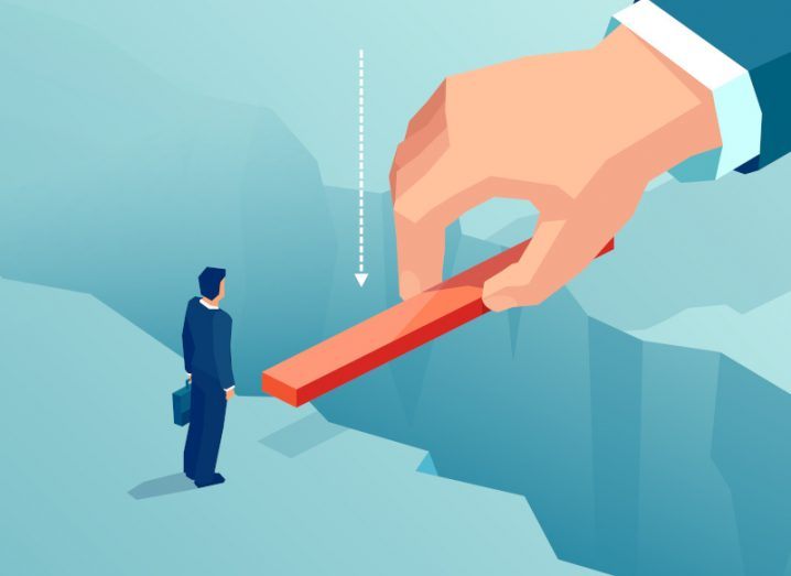 An illustration of a small businessman standing at the edge of a large crevice, wanting to get to the other side. A large hand is placing a plank of wood over the crevice to help him.