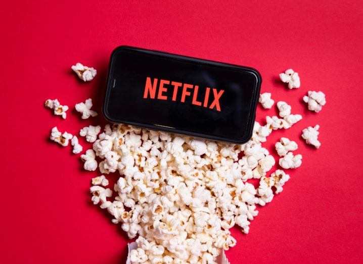 The Netflix logo on a smartphone on top of a pile of popcorn on a red surface.