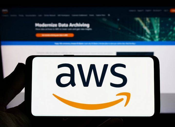The AWS logo on the screen of a smartphone, in front of a computer screen.
