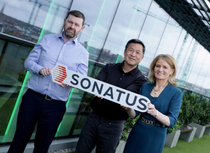 Two men and a woman holding a Sonatus sign in front of a building.