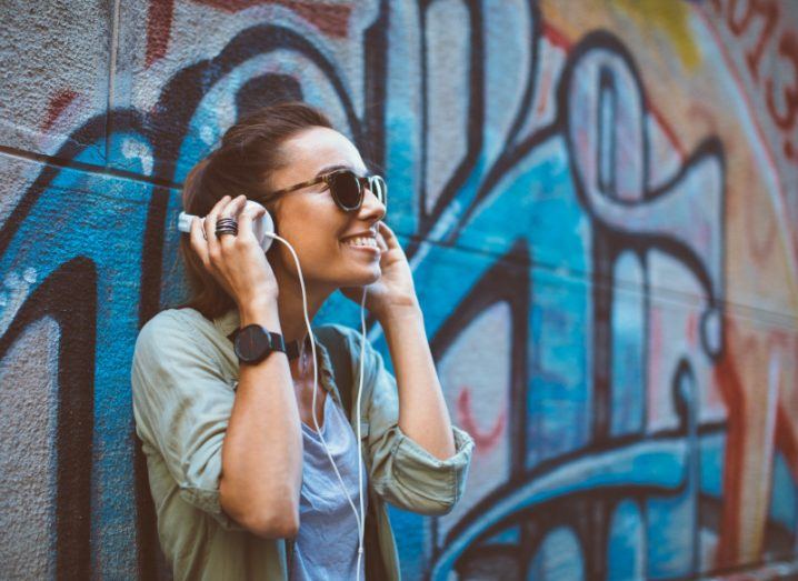 Woman listening to music on headphones while leaning against a wall with graffiti.
