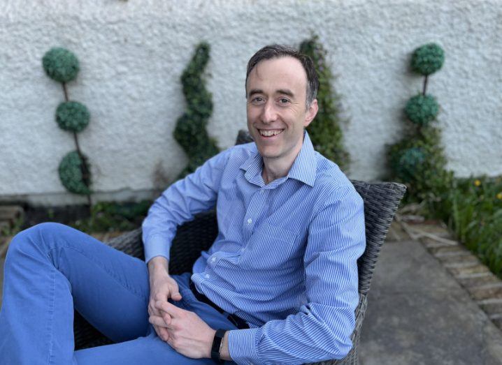 Barry Scott smiles at the camera, wearing a blue shirt and blue trousers. He is sitting on a patio chair in a garden.