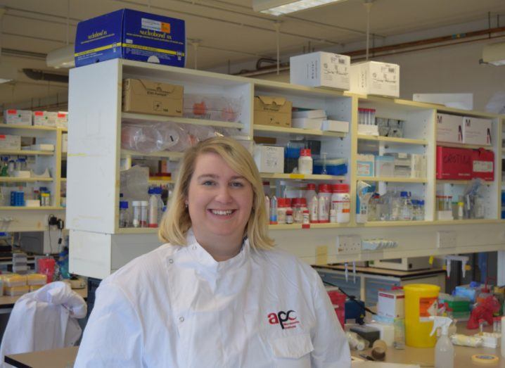 Dr Lily Keane in a fully-stocked lab wearing a white lab coat with APC Microbiome on it. She has bob-length blonde hair and smiles at the camera.