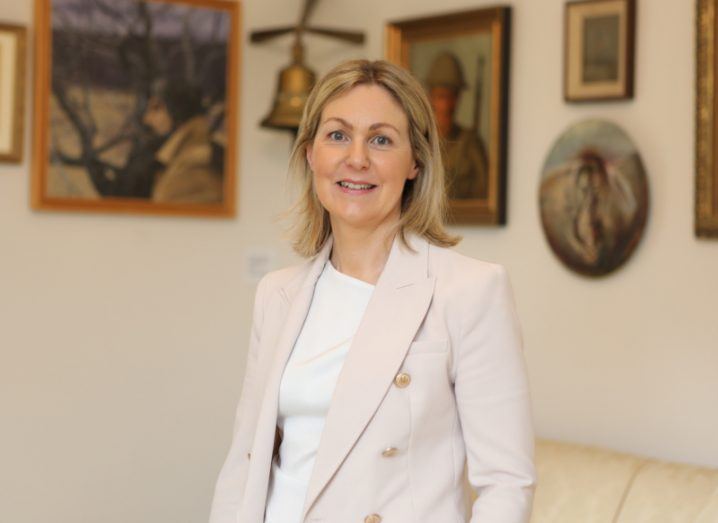 A woman wearing a cream-coloured suit jacket and a white blouse smiles at the camera in an ornate room filled with paintings. She is Professor Norelee Kennedy.