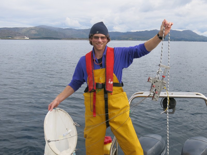Dr Damien Haberlin stands on a boat in fishing gear holding up a line in one hand and a net in the other for sampling plankton, with a grey sea and land visible behind him.