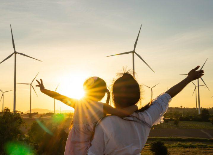 A woman stands holding a young child and the both face away from the camera towards a wind farm with arms outstretched and looking into a sunset.