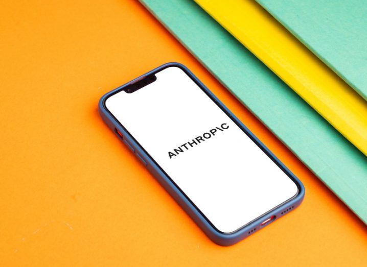 The Anthropic logo on a smartphone screen. The phone is laying on an orange surface.