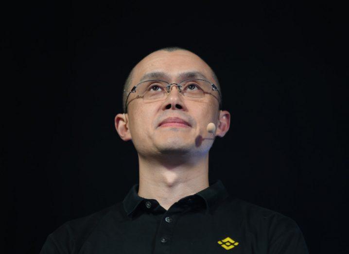 A headshot from below of Changpeng Zhao in a black t-shirt with a microphone taped to his cheek against a black background.